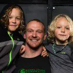 A South Lake Union chiropractor grins warmly, flanked by two joyful children, all clad in coordinated black and gray attire. Sports Medicine Northwest - West Seattle Chiropractic Clinic