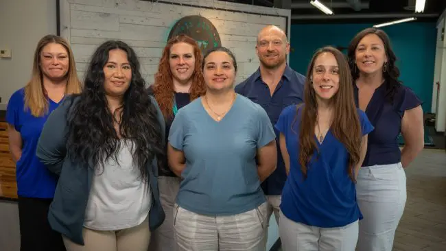 Seven people in business-casual attire stand indoors, smiling confidently at the camera, perhaps just after a meeting with a renowned West Seattle Chiropractor. Sports Medicine Northwest - West Seattle Chiropractic Clinic