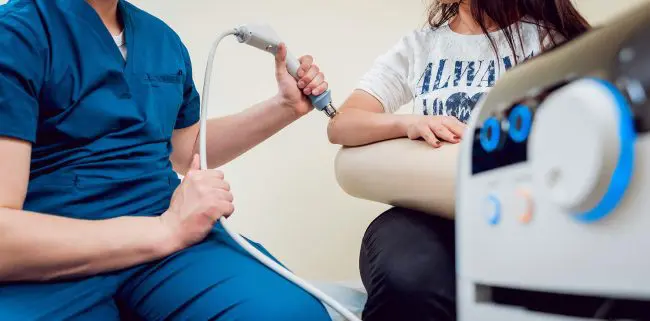 A medical professional in blue scrubs uses a handheld device on a patient's arm, administering shockwave therapy in a clinical setting in Seattle. Sports Medicine Northwest - West Seattle Chiropractic Clinic