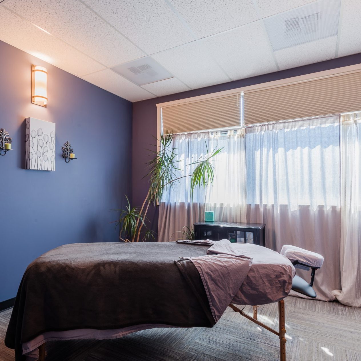 A massage room with a massage table, soft lighting, and blue walls. There is a window with curtains and a plant near the window. Sports Medicine Northwest - West Seattle Chiropractic Clinic