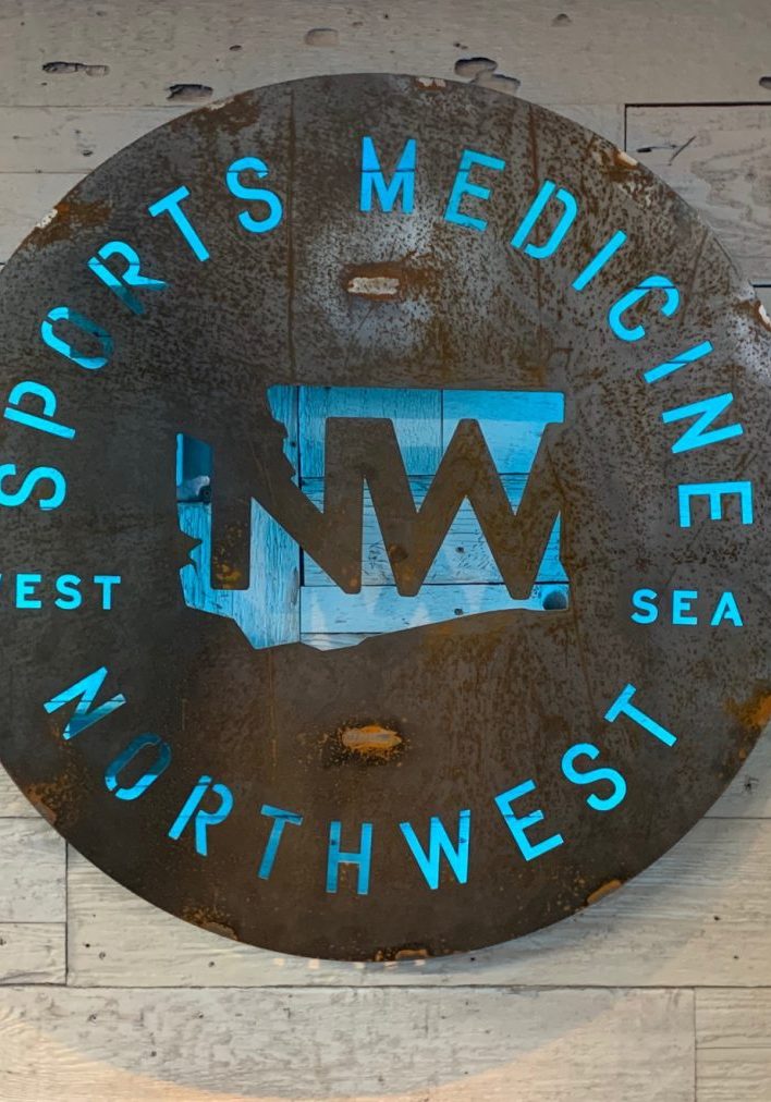 A round, metal sign on a wooden wall reads "Northwest Sports Medicine" with a cutout map and acronym "NW," emphasizing their expertise in corrective exercise. Sports Medicine Northwest - West Seattle Chiropractic Clinic