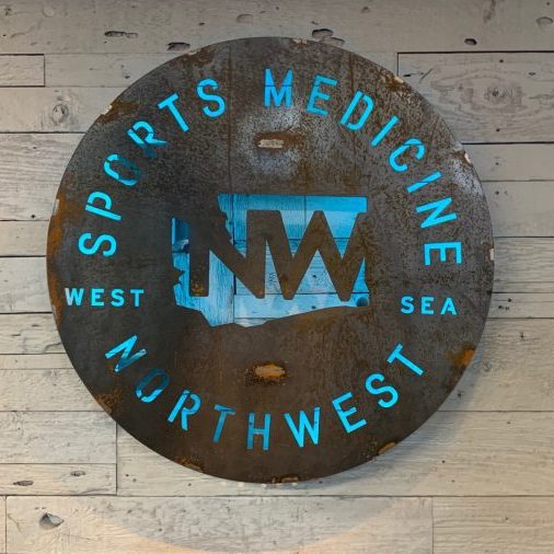 A round, metal sign on a wooden wall reads "Northwest Sports Medicine" with a cutout map and acronym "NW," emphasizing their expertise in corrective exercise. Sports Medicine Northwest - West Seattle Chiropractic Clinic