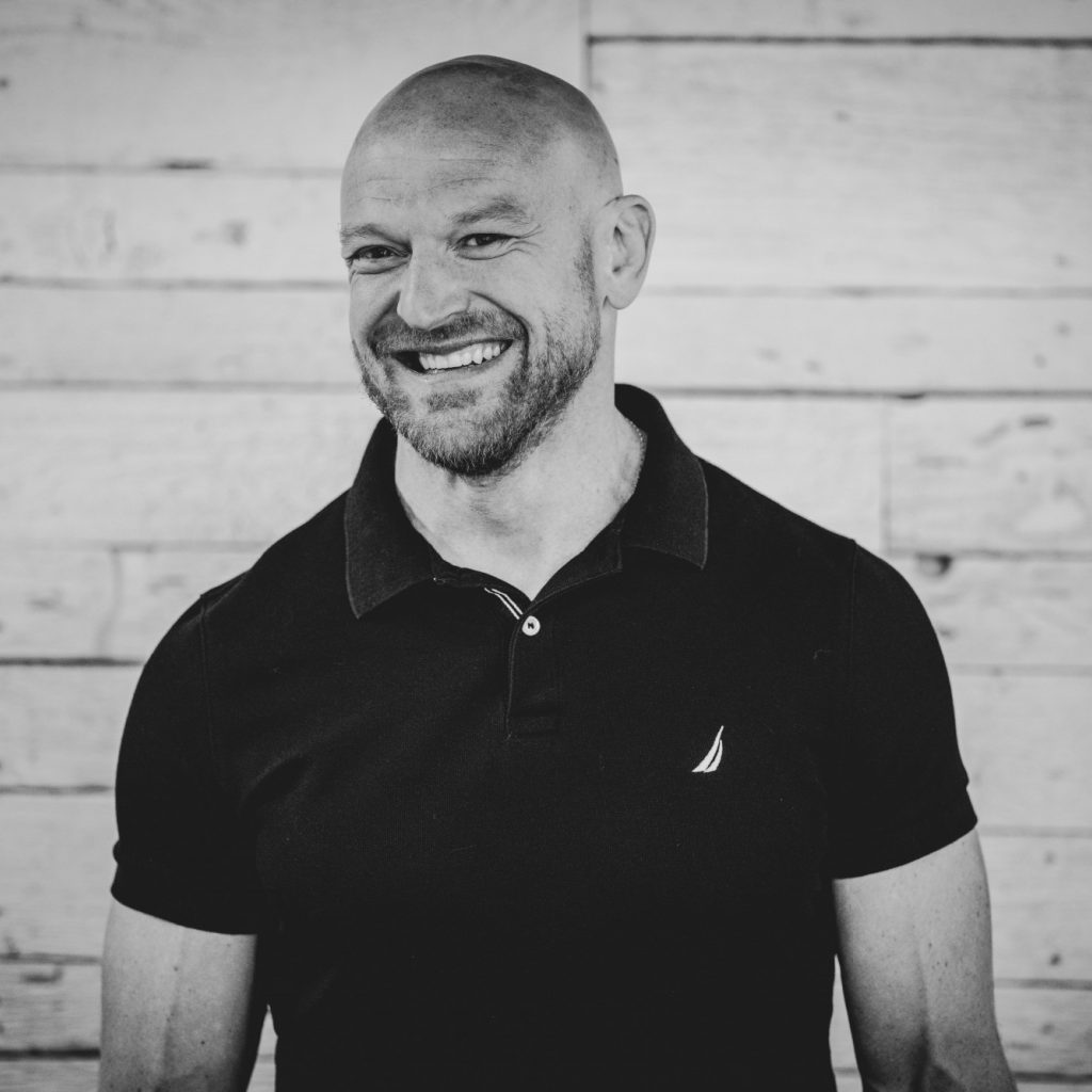 Skylar Pond in a black polo, your trusted West Seattle chiropractor, stands against a wooden plank wall. The image captures his professional yet approachable demeanor in classic black and white. Sports Medicine Northwest - West Seattle Chiropractic Clinic