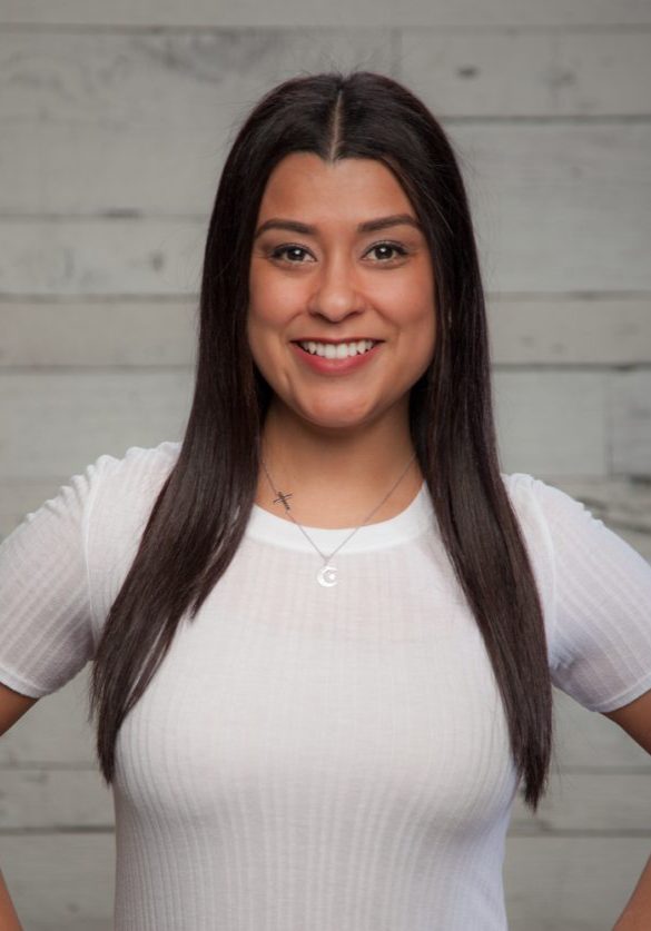 Meet Erica Cavazos, the Front Desk Manager in Seattle. She stands smiling with her long dark hair cascading over a crisp white shirt, set against a light wooden background. Sports Medicine Northwest - West Seattle Chiropractic Clinic