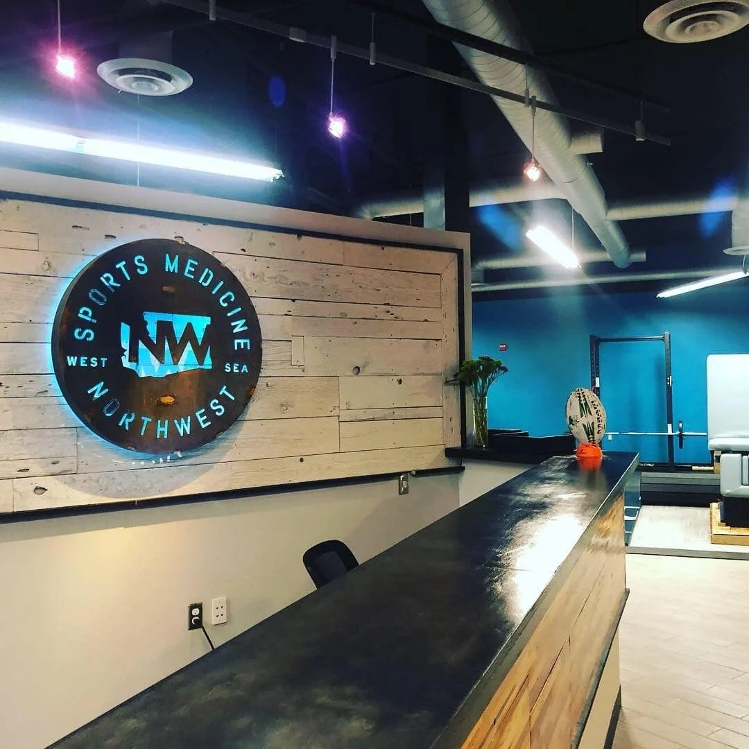 The reception area of Northwest Sports Medicine, a proud West Seattle Chiropractor, features a circular logo on a wooden wall, complemented by a sleek countertop and workout equipment subtly in the background. Sports Medicine Northwest - West Seattle Chiropractic Clinic