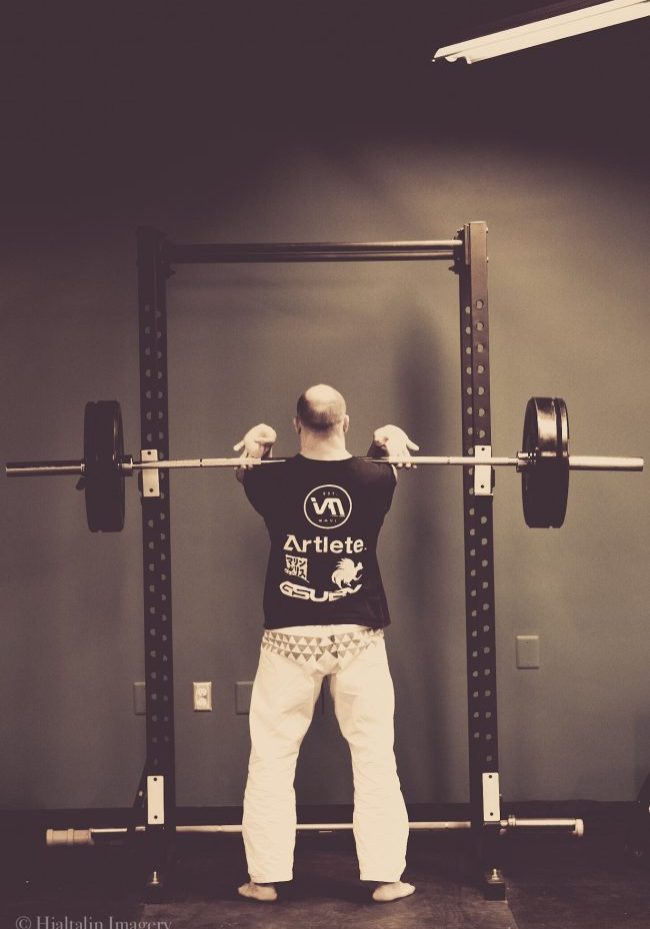 A person executing a barbell squat in the gym, focusing on proper form as part of their corrective exercise routine, dressed in a graphic t-shirt and white pants. Sports Medicine Northwest - West Seattle Chiropractic Clinic