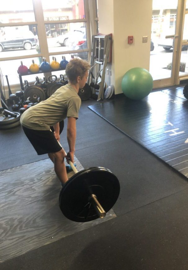 A person is performing a barbell deadlift in a gym, focusing on proper form as part of their corrective exercise routine, with various weights and equipment in the background. Sports Medicine Northwest - West Seattle Chiropractic Clinic