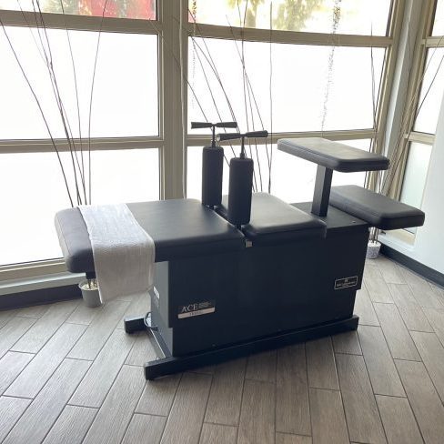 A black chiropractic adjustment table with a white towel draped over it is positioned on a tiled floor near large windows, ready for the renowned Ring Dinger technique in Seattle, Washington. Sports Medicine Northwest - West Seattle Chiropractic Clinic