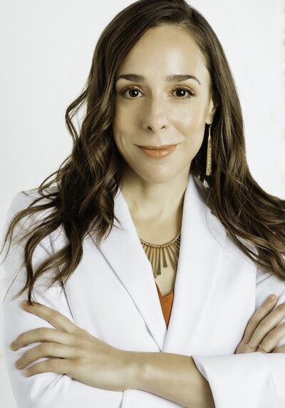 Dr. Lauren Procopio, a Naturopathic Doctor in Seattle, stands with her arms crossed against a neutral background. Her long brown hair cascades over a white coat, complemented by a subtle necklace that adds a touch of elegance to her professional aura. Sports Medicine Northwest - West Seattle Chiropractic Clinic