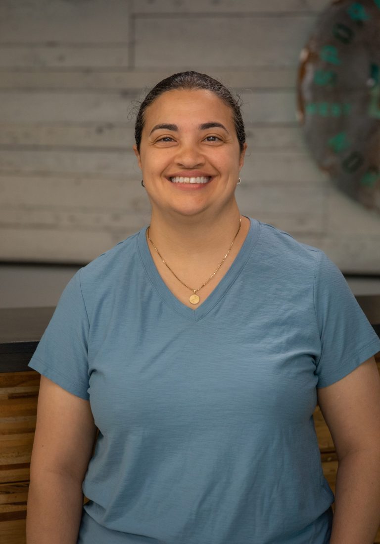 Dr. Jessica D’Auria, wearing a light blue shirt, smiles indoors against a wooden wall, with a decorative circular object behind her. Sports Medicine Northwest - West Seattle Chiropractic Clinic