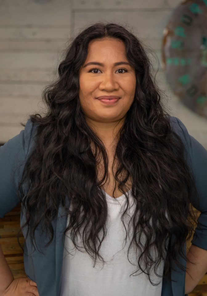 A person with long wavy hair and a blue blazer stands confidently with hands on hips, smiling at the camera. The wooden background and a blurry circular decor element provide a warm ambiance, embodying the spirit of teamwork across the ROW. Sports Medicine Northwest - West Seattle Chiropractic Clinic